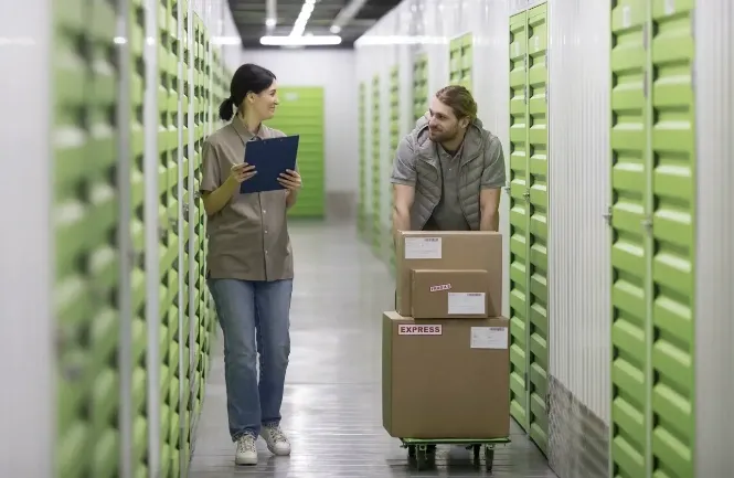 Customer moving boxes on a dolly while storage facility staff assists in a modern hallway with green self storage units